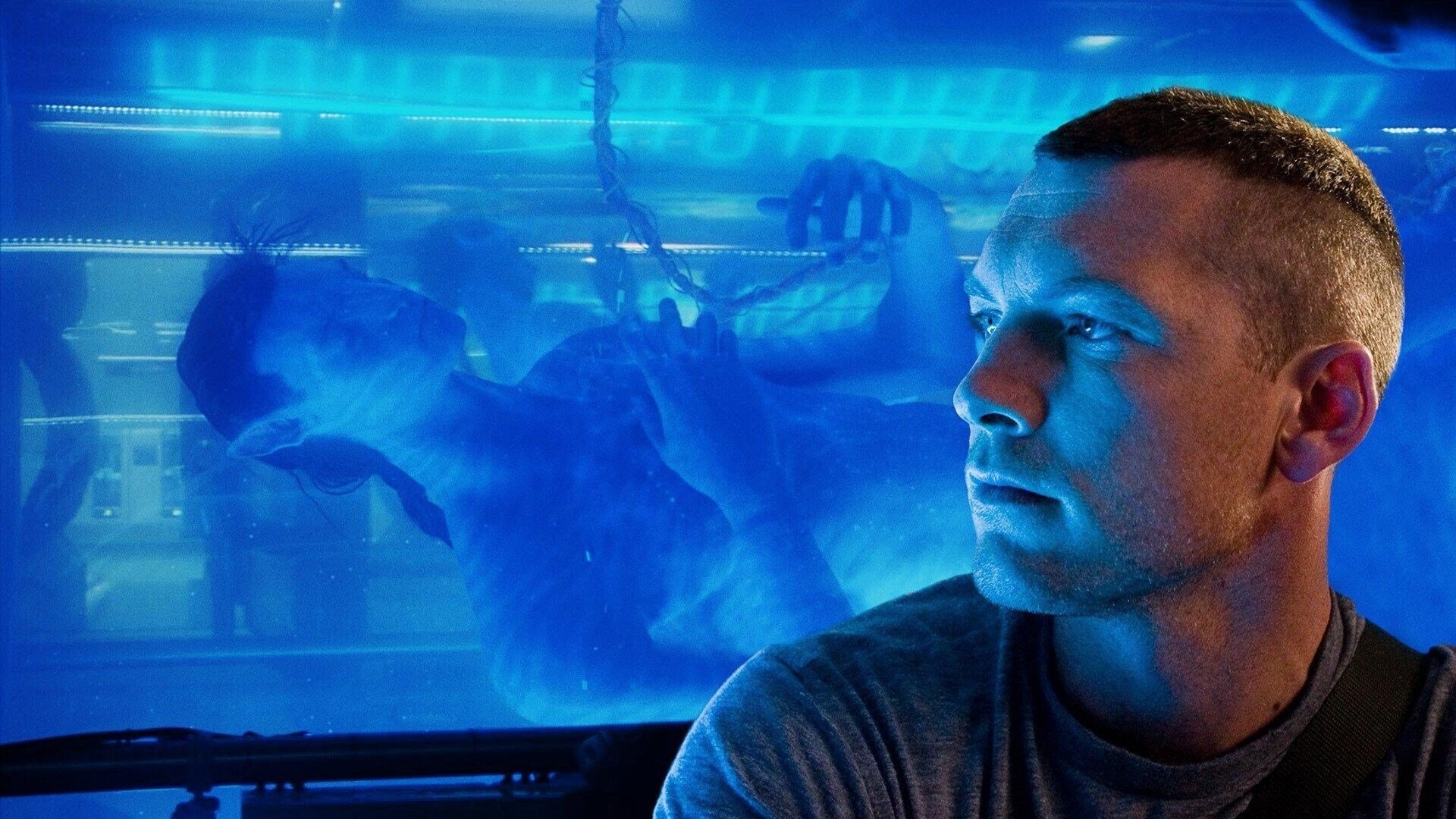 Homem humano observando avatar Na'vi flutuando em tanque de líquido azul iluminado em laboratório