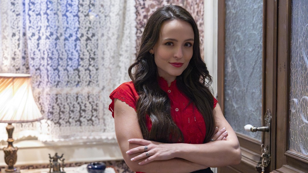 Mulher de blusa vermelha rendada e cabelo longo castanho em interior com cortinas brancas e luminária vintage