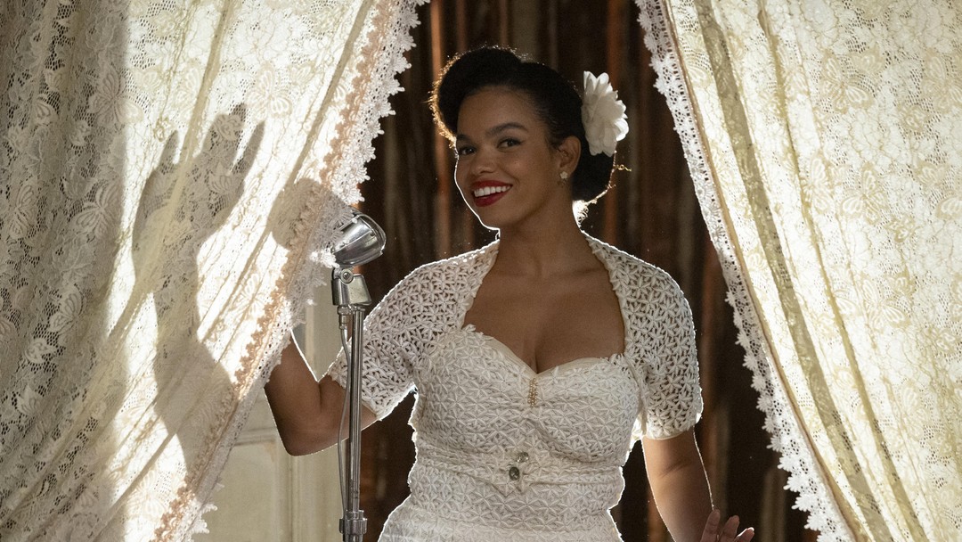 Mulher de cabelo preso e vestido branco rendado cantando em microfone vintage prateado entre cortinas de palco