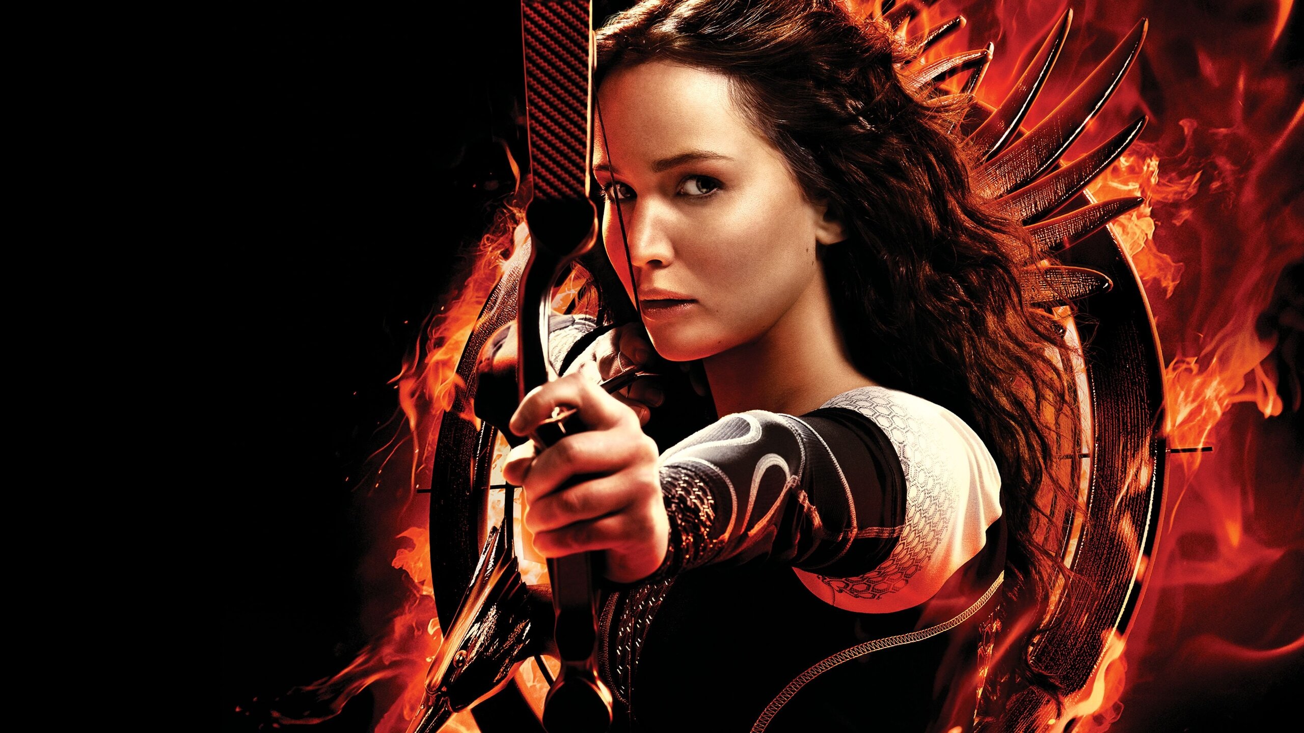 Mulher mirando arco e flecha diante de roda metálica em chamas com asas de mockingjay incandescentes ao fundo