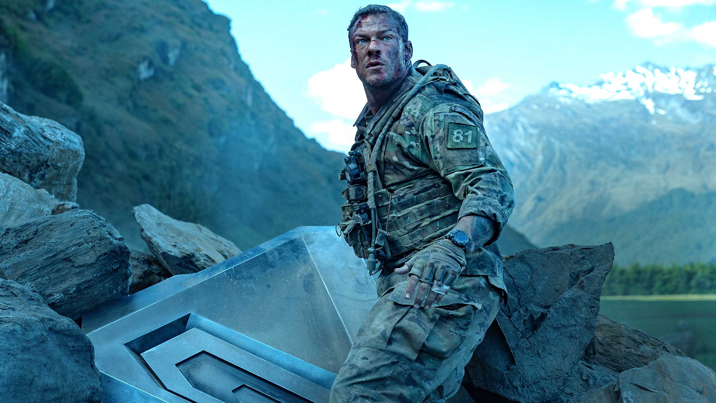 Alan Ritchson's 81 em pé ao lado de parte da máquina destruída e parecendo machucado em War Machine