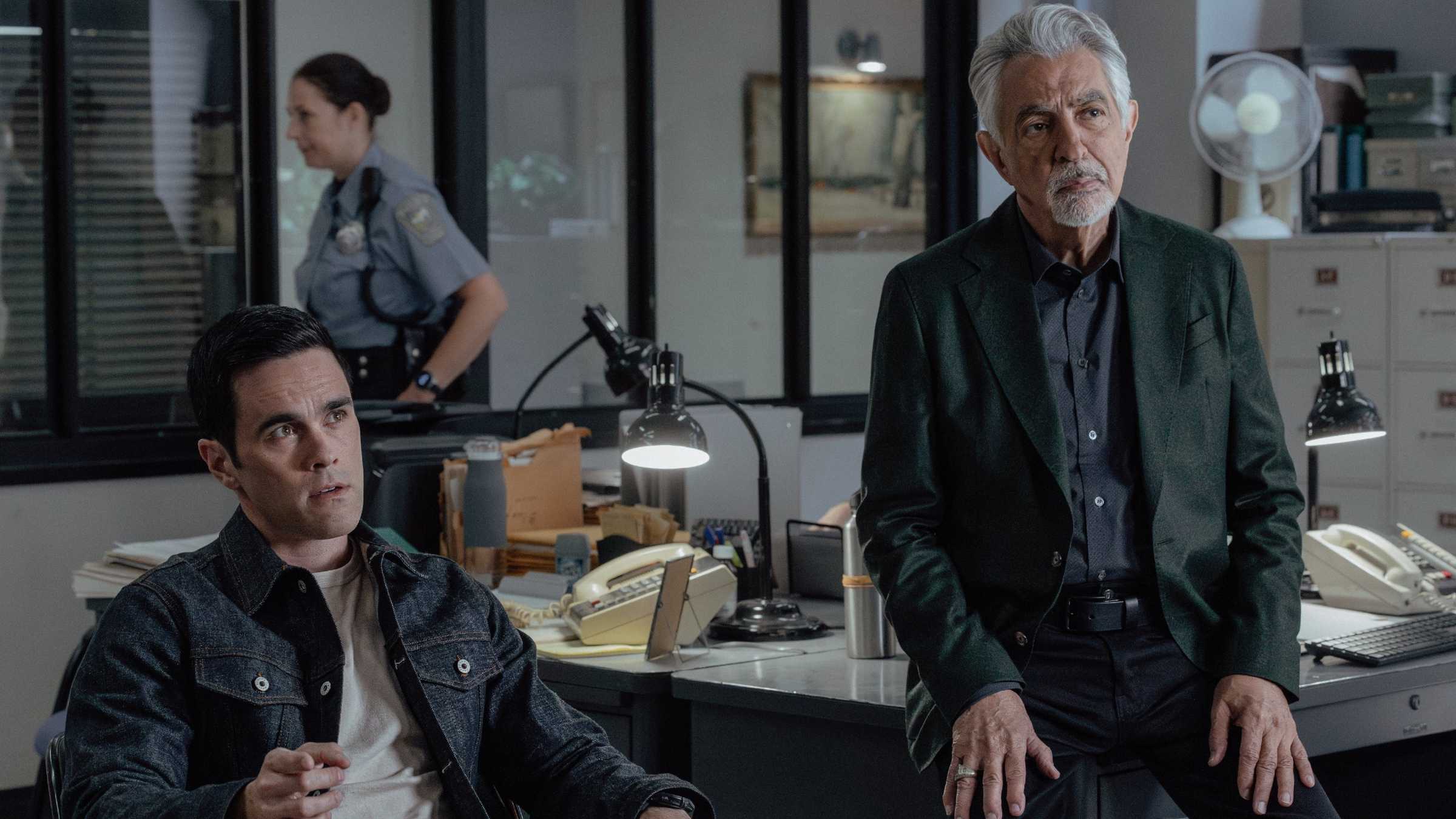 Cena de Criminal Minds com Tyler e Rossi sentados no escritório com um policial ao fundo.