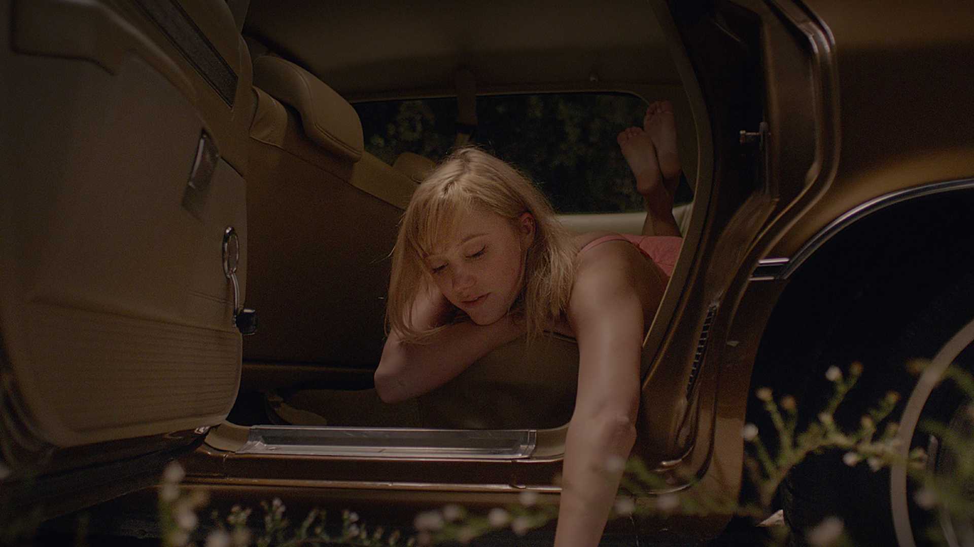 Maika Monroe como Jay deitada no banco de trás de um carro, inclinando-se para fora da porta e tocando a grama em It Follows