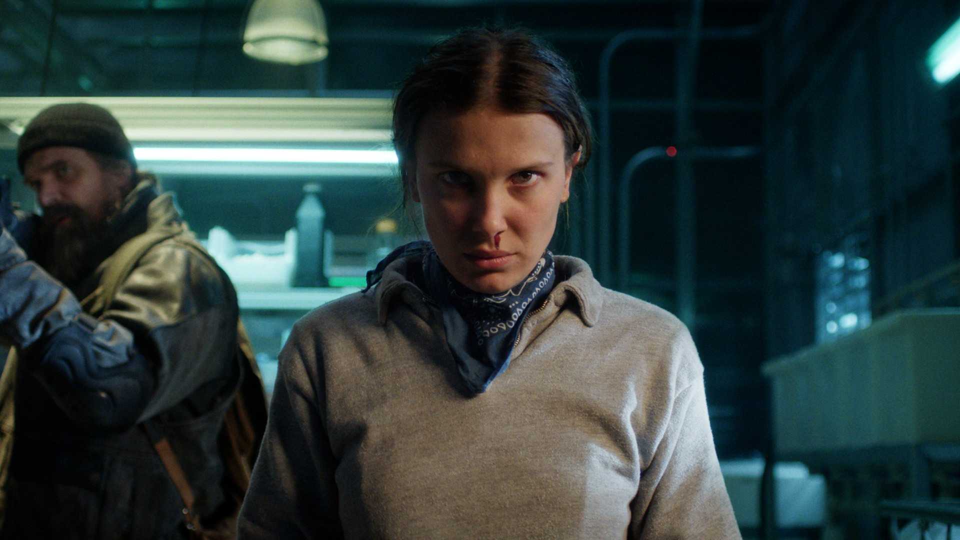 Millie Bobby Brown como Eleven em Stranger Things temporada 5, David Harbour como Hopper carregando uma arma ao fundo