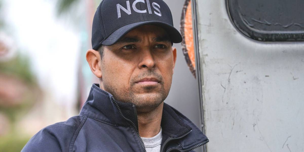 Nick Torres usando um boné do NCIS e um corta-vento azul, parado ao lado de uma van na série de TV NCIS