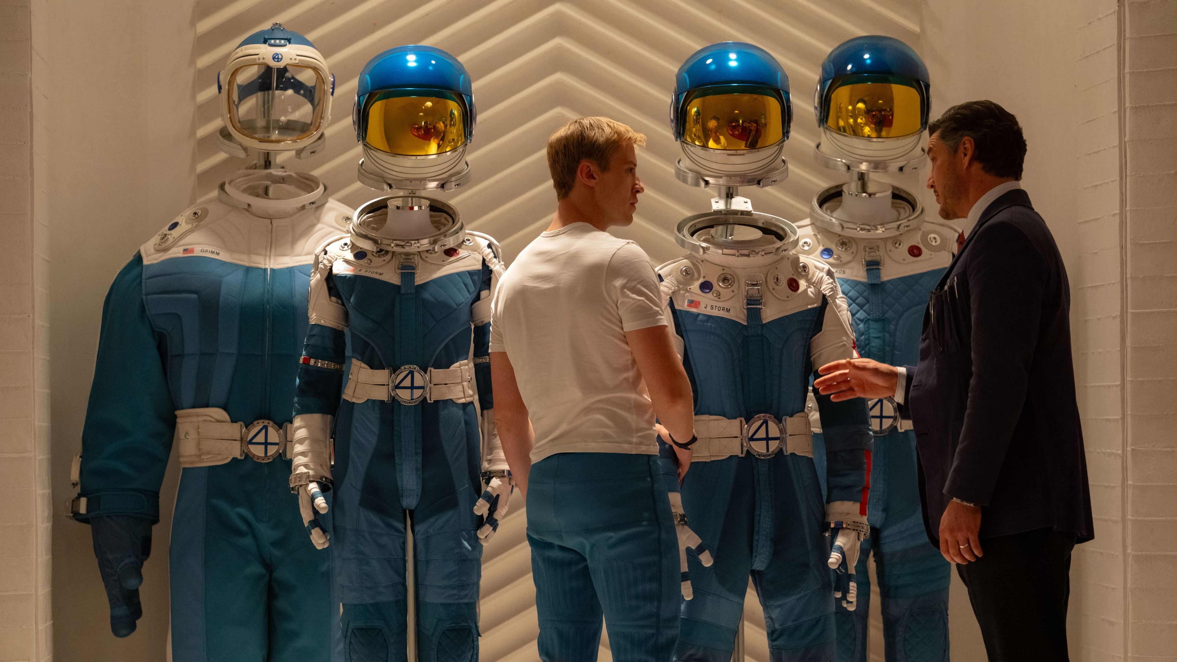 Quatro astronautas em uniformes brancos e azuis com capacetes dourados em instalação futurista durante preparação para missão