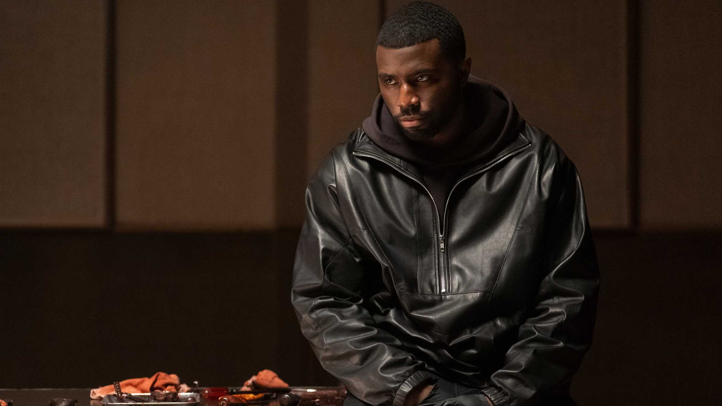 Coltrane de Y'lan Noel sentado em uma mesa com armas sobre ela em Nemesis